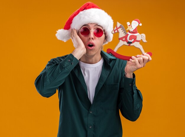 Bezorgde jonge blonde man met kerstmuts en bril met santa op schommelpaard beeldje kijken camera houden hand op gezicht geïsoleerd op oranje achtergrond