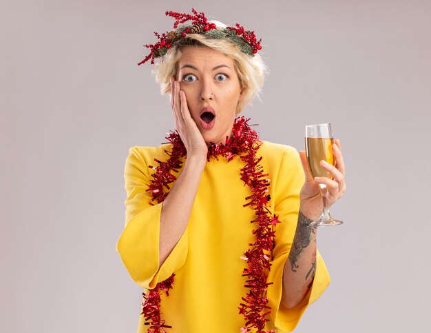 Bezorgde jonge blonde vrouw draagt ?? kerst hoofd krans en klatergoud slinger rond de nek met glas champagne kijken camera hand houden op gezicht geïsoleerd op witte achtergrond