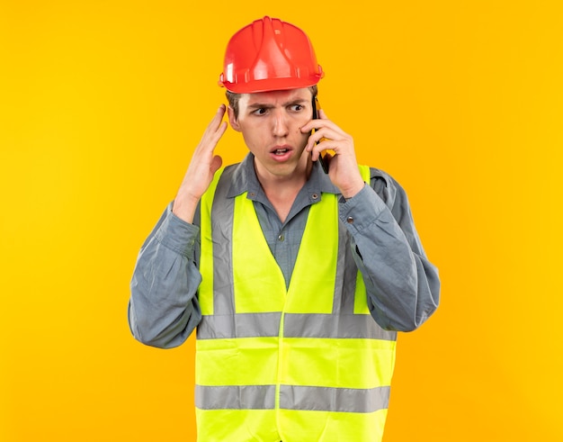 Bezorgde jonge bouwer man in uniform spreekt op telefoon geïsoleerd op gele muur