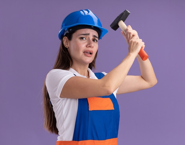 Bezorgde jonge bouwvrouwen in uniform die hamer opheffen die op paarse muur wordt geïsoleerd