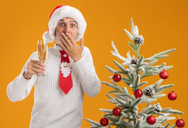 bezorgde jonge knappe kerel met een kerstmuts en een stropdas van de kerstman die in de buurt van een versierde kerstboom staat met een glas champagne met de hand op de mond en kijkt geïsoleerd op een oranje muur