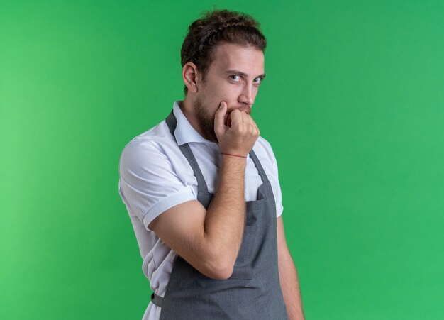 Bezorgde jonge mannelijke kapper met uniform bijt nagels geïsoleerd op groene muur green