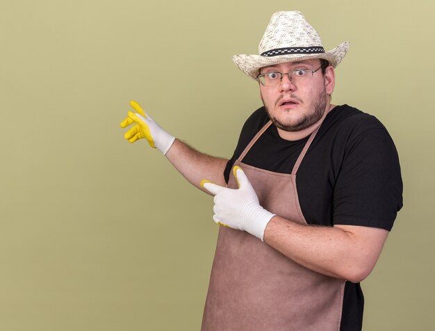 Bezorgde jonge mannelijke tuinman met tuinhoed en handschoenen wijst naar achter geïsoleerd op olijfgroene muur met kopieerruimte copy