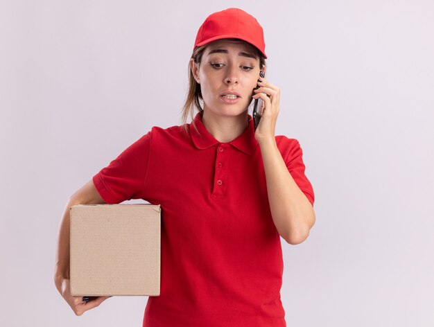 Bezorgde jonge mooie leveringsvrouw in uniform houdt kartonnen doos en praat over telefoon geïsoleerd op een witte muur