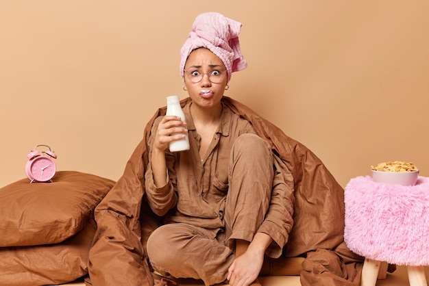 Bezorgde jonge vrouw draagt gewikkelde handdoek op hoofd en pyjama drinkt verse melk uit fles heeft snel ontbijt poses op bed onder deken geïsoleerd over bruine achtergrond Ontbijt tijd concept