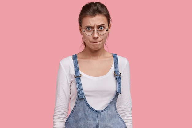 Gratis foto bezorgde jonge vrouw drukt lippen met verwarde gezichtsuitdrukking, probeert een uitweg te vinden, heeft een verbaasde blik, draagt een ronde grote bril
