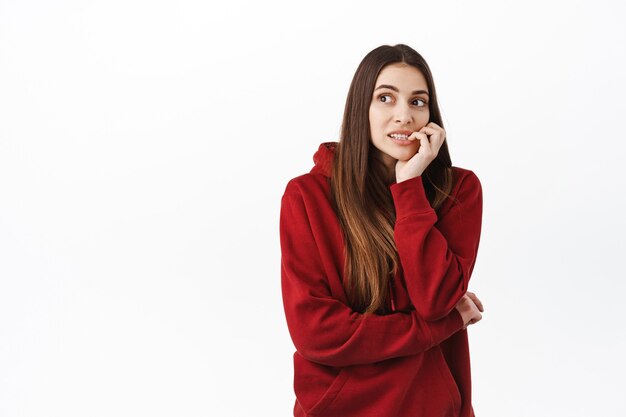 Bezorgde jonge vrouw met lang haar, vingernagels bijtend en peinzend opzij kijkend, nerveus denkend, moeilijke keuze makend, nadenkend tegen de witte muur staan