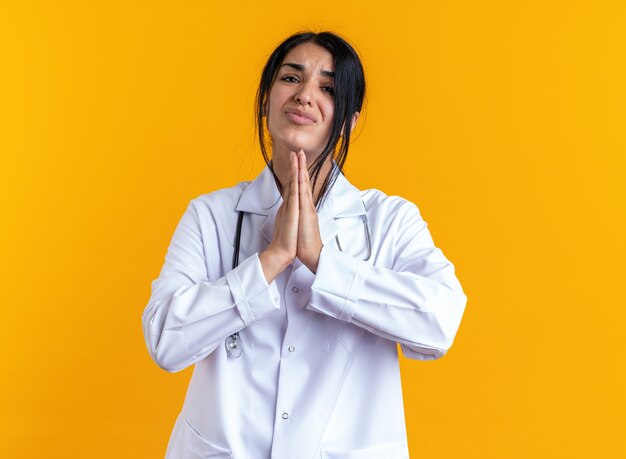 Bezorgde jonge vrouwelijke arts die een medisch gewaad draagt met een stethoscoop die een gebedsgebaar toont dat op een gele muur is geïsoleerd