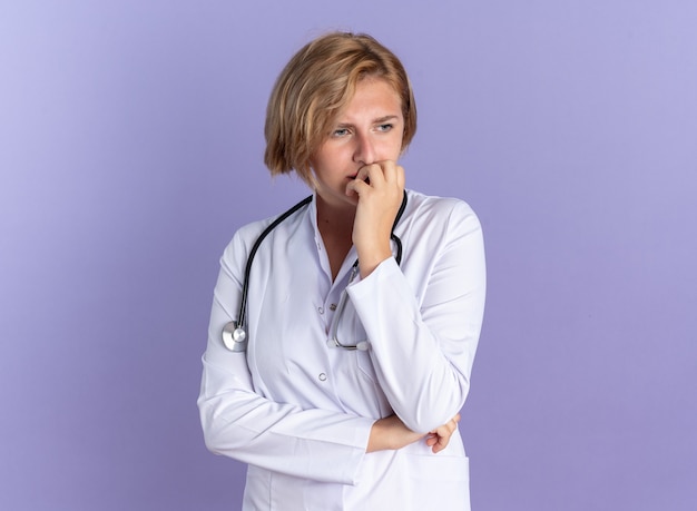 Bezorgde jonge vrouwelijke arts die medische mantel draagt met stethoscoop bijt nagels geïsoleerd op blauwe muur