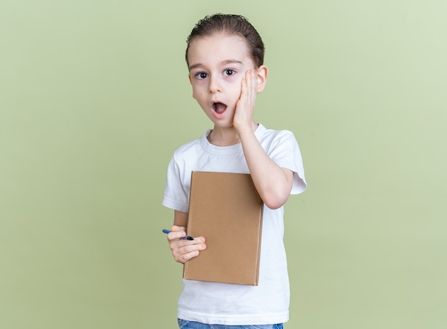 Bezorgde kleine jongen die hand op zijn gezicht houdt met boek en kijkt naar camera geïsoleerd op olijfgroene muur met kopieerruimte