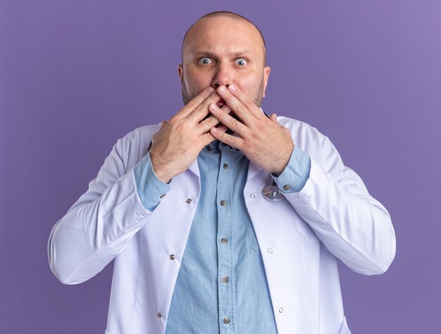 Bezorgde mannelijke arts van middelbare leeftijd die een medische mantel en een stethoscoop draagt, die de handen op de mond houdt, geïsoleerd op de paarse muur