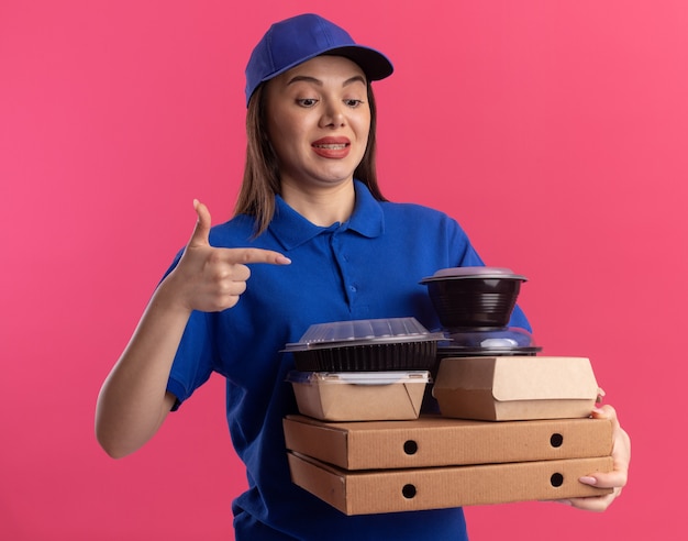 Bezorgde mooie bezorger in uniform houdt en wijst naar voedselpakket en containers op pizzadozen op roze