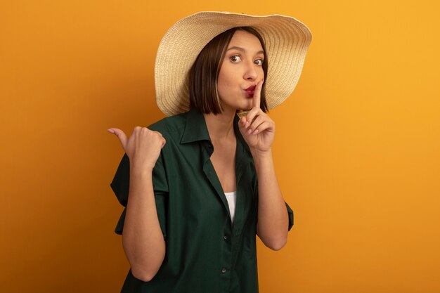 Bezorgde mooie vrouw die met strandhoed stiltegebaar doet en wijst aan kant die op oranje muur wordt geïsoleerd