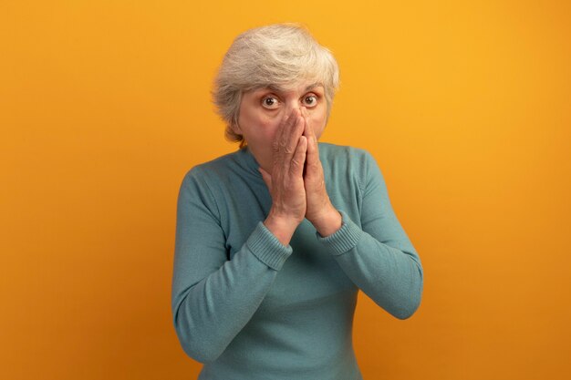 Bezorgde oude vrouw met een blauwe coltrui die naar de voorkant kijkt en de handen bij elkaar houdt op de mond geïsoleerd op een oranje muur met kopieerruimte