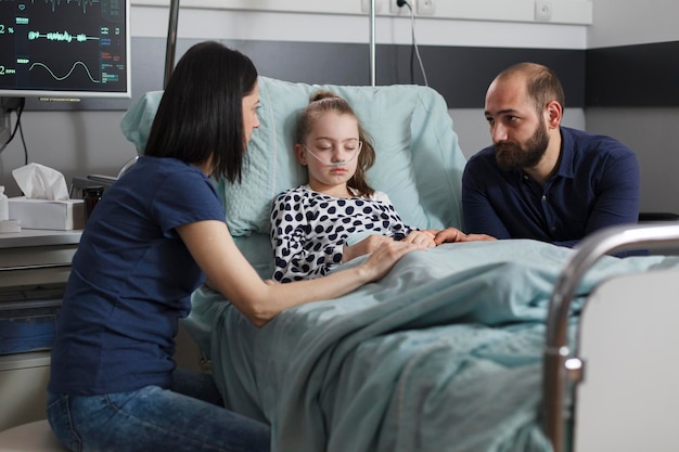 Bezorgde ouders zitten naast een zieke dochter die in het ziekenhuis is opgenomen in een pediatrische zorginstelling. Slapend ziek meisje getroost door zorgzame moeder en vader terwijl ze in het bed van de patiënt rust.