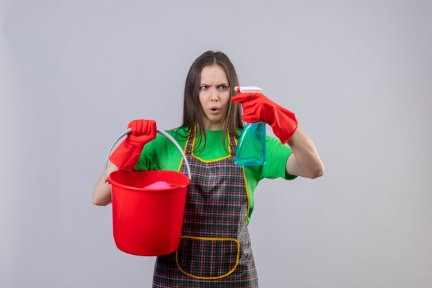 Bezorgde schoonmaak jonge vrouw, gekleed in uniform in rode handschoenen met emmer op zoek naar schoonmaak spray op haar hand op geïsoleerde witte muur