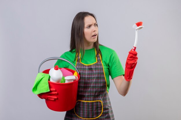 Bezorgde schoonmaakster die uniform in rode handschoenen draagt die schoonmaakgereedschap houdt op zoek naar schoonmaakborstel op haar hand op geïsoleerde witte muur