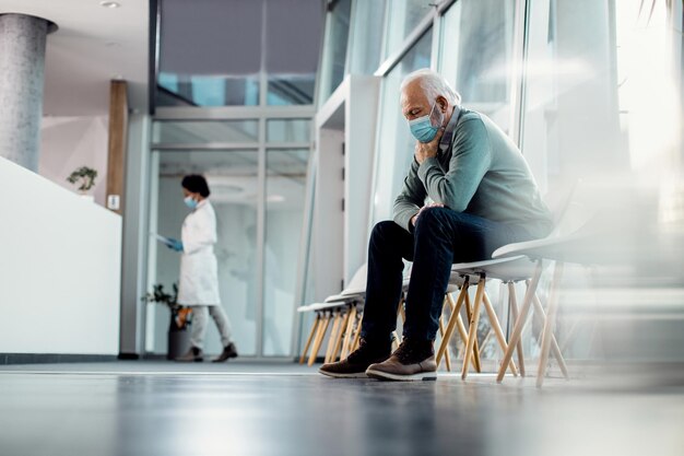 Bezorgde senior man die aan iets denkt terwijl hij in de wachtkamer van het ziekenhuis zit tijdens een pandemie van het coronavirus