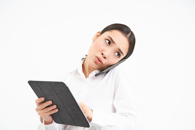 Bezorgde verkoopster Aziatische kantoordame die tijdens het spreken naar een digitale tablet kijkt die de telefoon op de schouder houdt