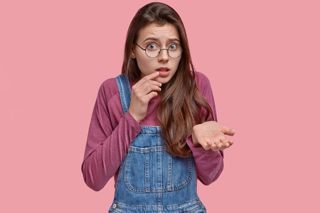 Bezorgde verontwaardigde vrouw steekt handpalm op, kijkt gefrustreerd, draagt een ronde bril, denim tuinbroek