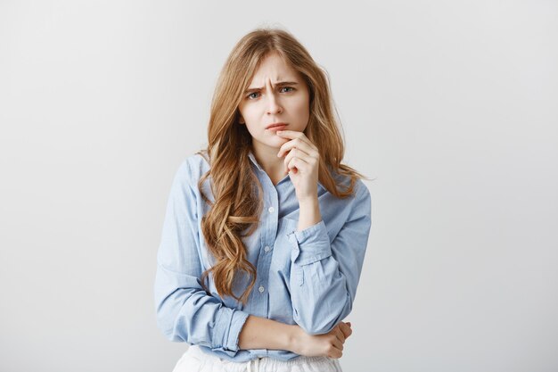 Bezorgde vriend die het probleem hoort. Portret van aantrekkelijke blanke vrouw met blond haar en krullen die hand op de lip houden, fronsend, bezorgd en serieus terwijl ze een oplossing bedenken