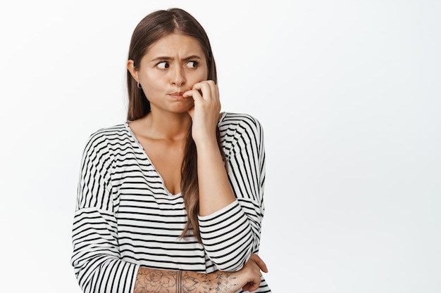 Bezorgde vrouw die vingers bijt en opzij kijkt, nerveus staat, zich zorgen maakt over iets, over een witte achtergrond staat