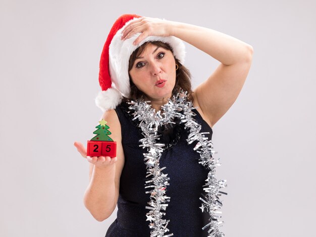 Bezorgde vrouw van middelbare leeftijd met een kerstmuts en een klatergoudslinger om de nek met kerstboomspeelgoed met datum die naar de camera kijkt en de hand op het hoofd houdt geïsoleerd op een witte achtergrond met kopieerruimte