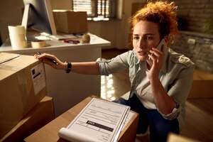 Bezorgde vrouwelijke koerier die via de mobiele telefoon communiceert tijdens het werken met bezorgpakketten op kantoor
