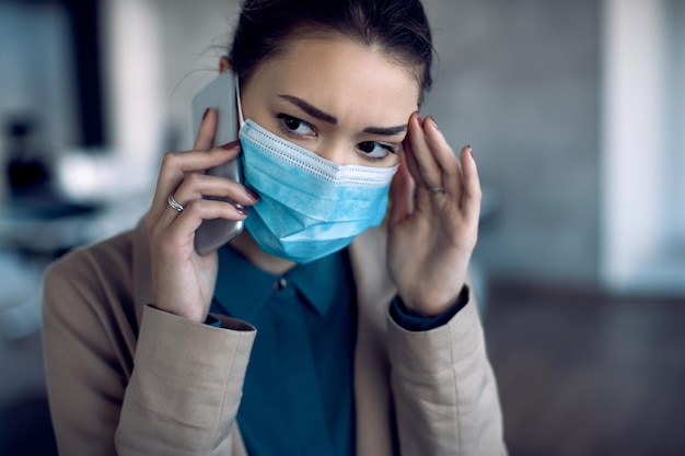 Bezorgde zakenvrouw die via mobiele telefoon communiceert terwijl ze een beschermend gezichtsmasker op kantoor draagt