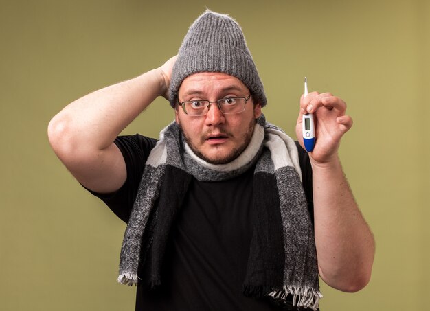Bezorgde zieke man van middelbare leeftijd met een wintermuts en sjaal die een thermometer vasthoudt en de hand op het hoofd legt