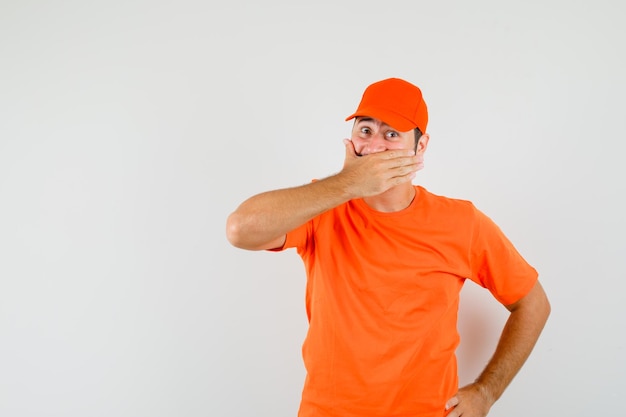 Bezorger houdt hand op mond in oranje t-shirt, pet en kijkt opgewonden. vooraanzicht.
