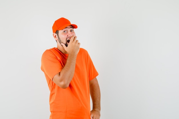 Bezorger houdt hand op mond in oranje t-shirt, pet en ziet er gelukkig uit, vooraanzicht.