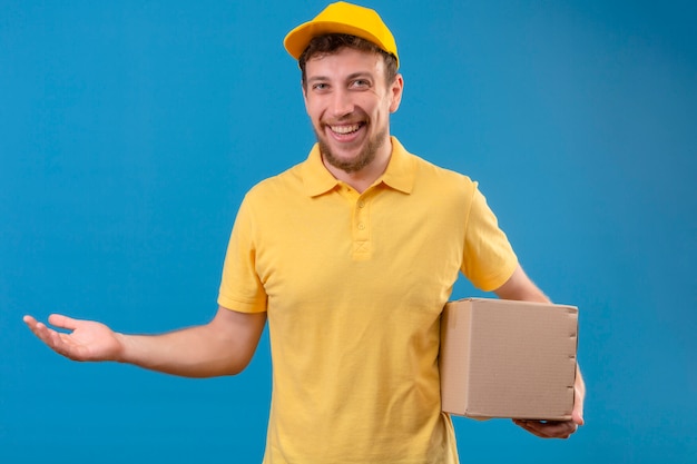 bezorger in geel poloshirt en pet bedrijf box pakket presenteren met palm oh zijn hand glimlachend vriendelijke staande op geïsoleerde blauw