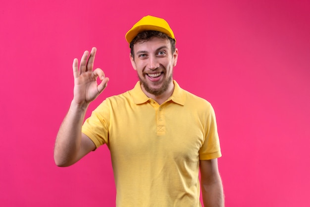 bezorger in geel poloshirt en pet kijken camera glimlachen vriendelijk doen ok teken staande op geïsoleerde roze