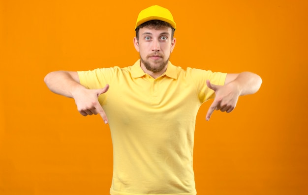 bezorger in geel poloshirt en pet kijken camera wijzend met vinger naar beneden kijken zelfverzekerd staande op oranje