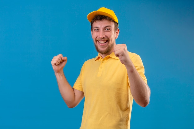 bezorger in geel poloshirt en pet kijkt opgewonden verheugd over zijn succes en overwinning zijn vuisten balancerend van vreugde blij om zijn doel en doelen te bereiken staande op geïsoleerd blauw