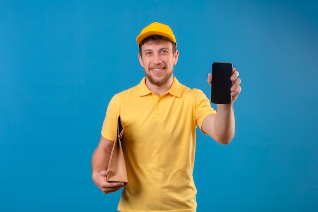 Bezorger in geel poloshirt en pet met papieren pakket en smartphone glimlachend vriendelijk met blij gezicht staande op blauw tonen