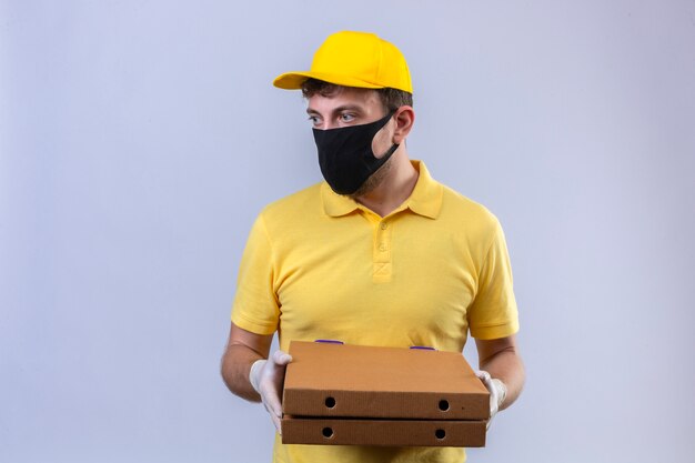 bezorger in geel poloshirt en pet met zwart beschermend masker houden pizzadozen opzij kijken met ernstig gezicht staande op geïsoleerde wit