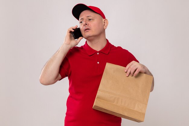 Bezorger in rood uniform en pet met papieren pakket die er zelfverzekerd uitziet terwijl hij op een mobiele telefoon praat die over een witte achtergrond staat