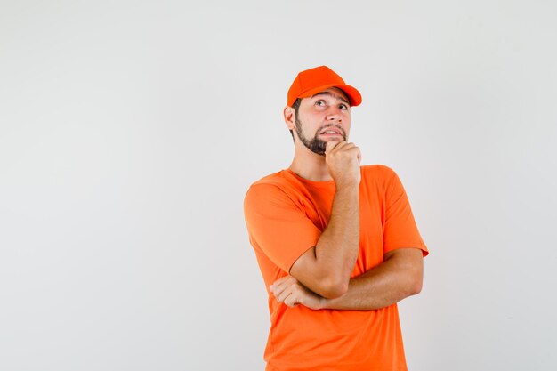 Bezorger kijkt op terwijl hij zijn kin vasthoudt in een oranje t-shirt, pet en peinzend kijkt. vooraanzicht.