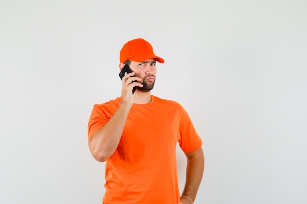 Bezorger praten op mobiele telefoon in oranje t-shirt, pet en peinzend kijken. vooraanzicht.