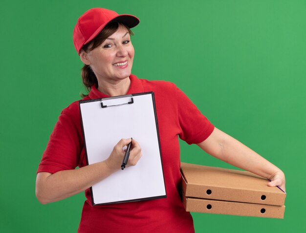 Bezorger van middelbare leeftijd in rood uniform en pet met pizzadozen en klembord met blanco pagina's en pen op zoek naar voorzijde lachend vragen om handtekening staande over groene muur