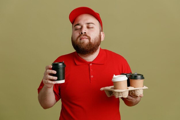 Bezorger werknemer in rode dop blanco t-shirt uniform met koffiekopjes blij en tevreden staande over groene achtergrond