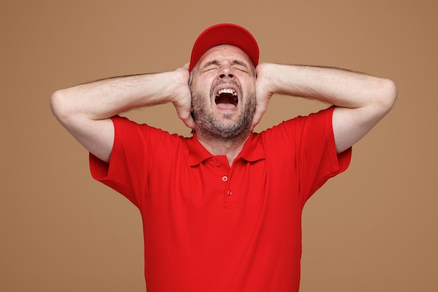 Bezorger werknemer in rode dop blanco tshirt uniform boos en gefrustreerd schreeuwen met geërgerde uitdrukking hand in hand op zijn hoofd staande over bruine achtergrond