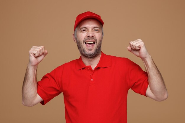 Bezorger werknemer in rode dop leeg t-shirt uniform kijken camera gek blij en opgewonden verhogen gebalde vuisten zich verheugen over zijn succes permanent over bruine achtergrond