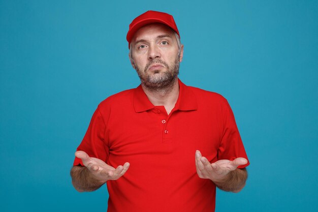Bezorger werknemer in rode dop leeg t-shirt uniform kijken naar camera verward opheffende armen twijfelend over blauwe achtergrond