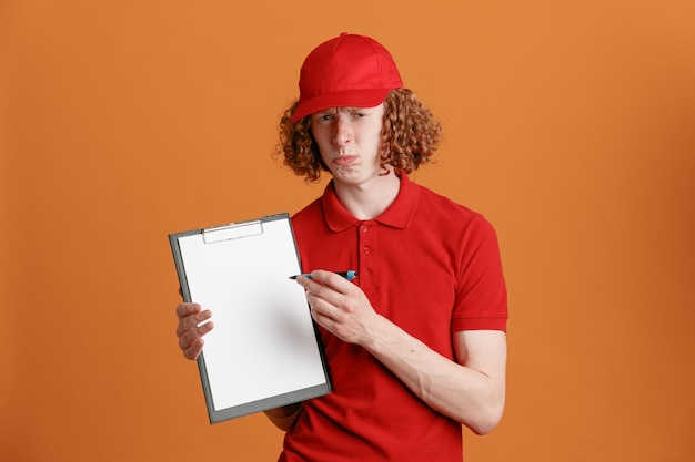 Bezorger werknemer in rode dop leeg t-shirt uniform met klembord met pen kijkend naar camera met droevige uitdrukking wachtend op handtekening staande over oranje achtergrond