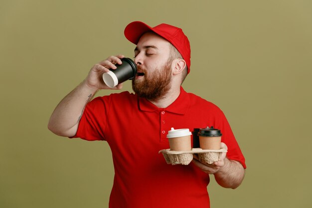 Bezorger werknemer in rode dop leeg t-shirt uniform met koffiekopjes drinkend ziet er gelukkig en positief uit over groene achtergrond