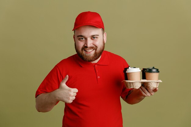 Bezorger werknemer in rode dop leeg t-shirt uniform met koffiekopjes kijkend naar camera gelukkig en vrolijk glimlachen duim opdagen staande over groene achtergrond