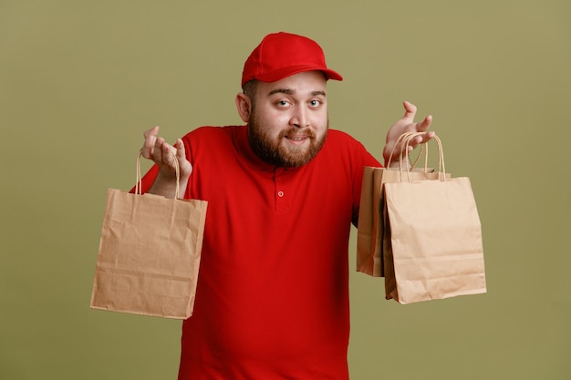 Bezorger werknemer in rode dop leeg t-shirt uniform met papieren zakken kijkend naar camera gelukkig en positief glimlachend vriendelijk staande over groene achtergrond